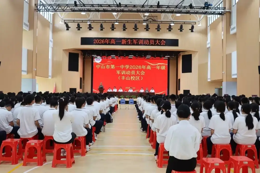 Alunos participando de uma assembleia escolar na Escola Secundária nº 1 de Zhongshan, com um grande telão de LED exibindo informações sobre o evento de treinamento militar para calouros de 2026.